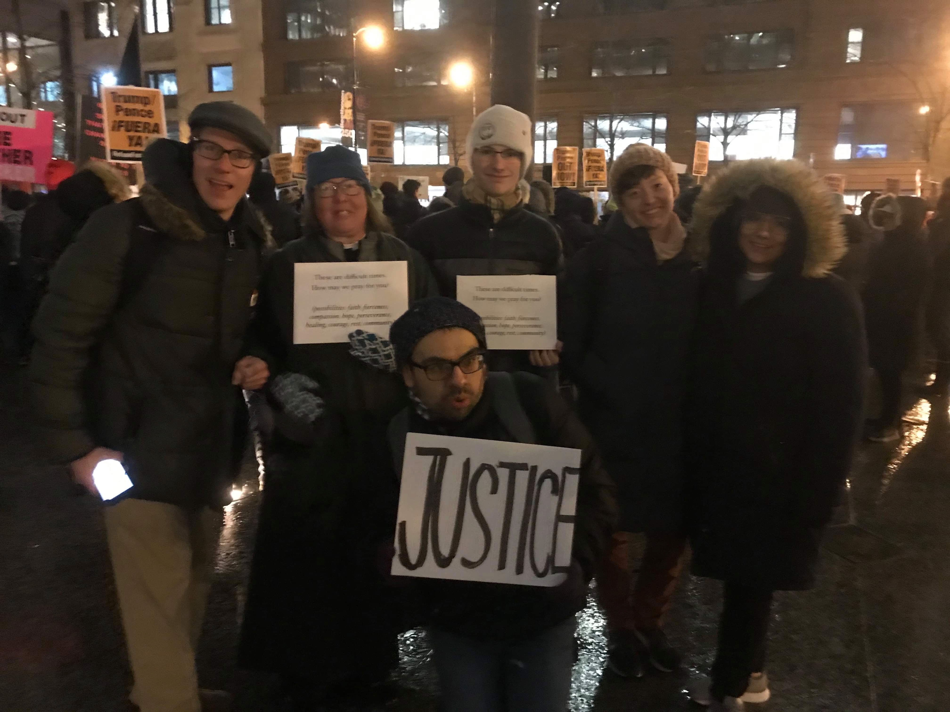 Brent House folks dressed in hats and winter coats  (but no masks—this was before the pandemic) offer prayers and support at a protest. In the middle of the group, someone holds a sign that says “JUSTICE” in big, black letters.